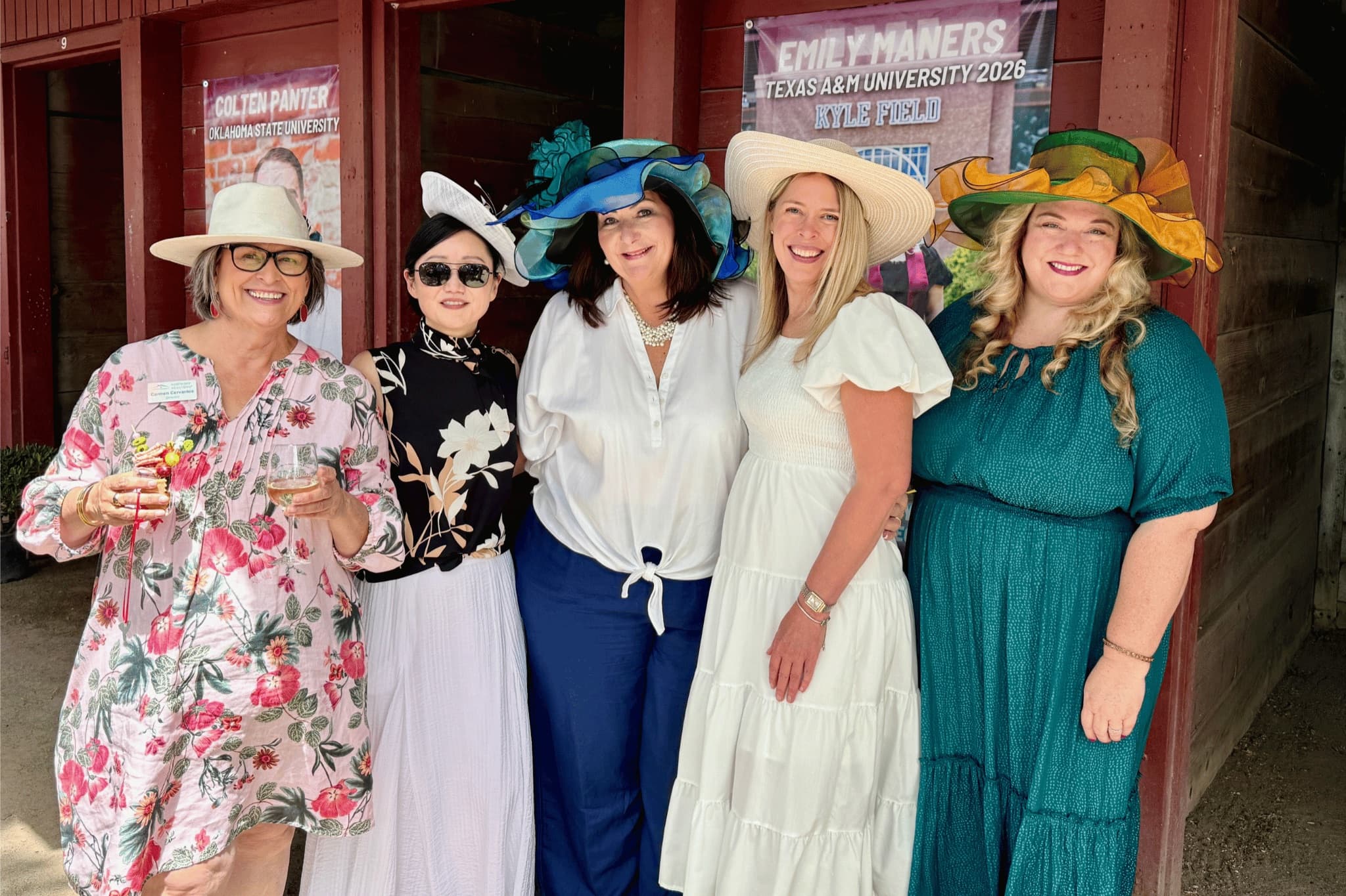 Five NORBAR members in wide-brimmed derby hats smiling outside a red barn at a charity race day