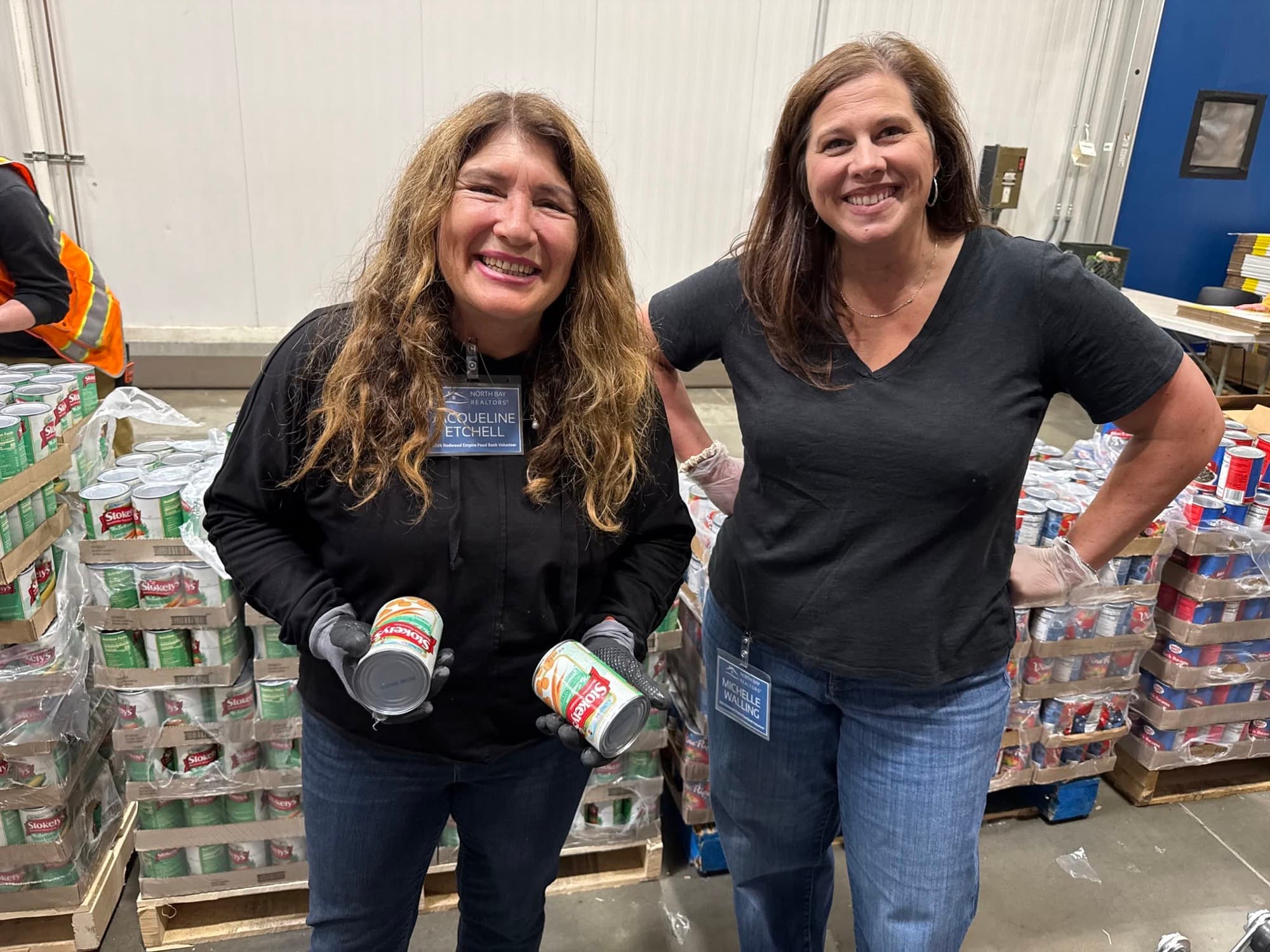 Two NORBAR volunteers in name badges holding canned goods among pallets at the Redwood Empire Food Bank