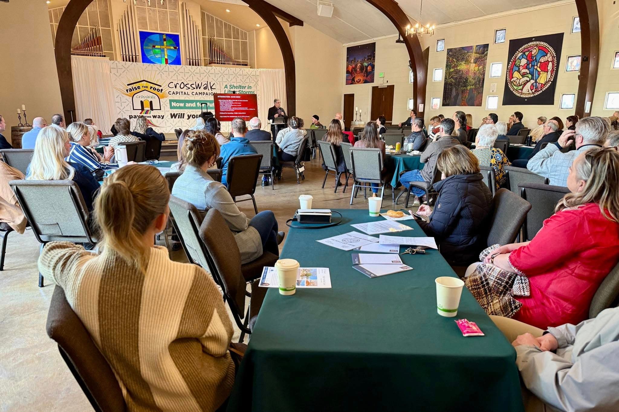 NORBAR members filling a hall for a chapter meeting, seated around cloth-covered tables with handouts and coffee while a speaker presents from a lectern at the front