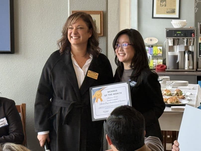 Two women smiling at a chamber meeting while holding a framed-style Member of the Month certificate.