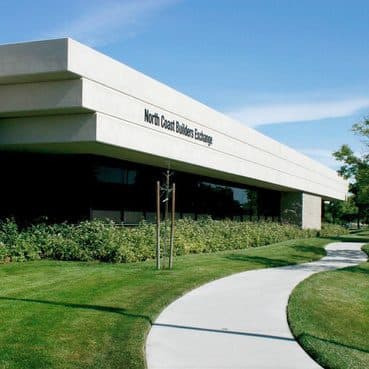 Exterior of the low white North Coast Builders Exchange building, with lawn and a curving walkway leading to the entrance.