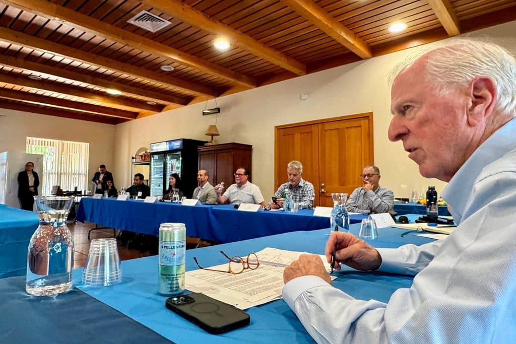 People seated along blue-draped tables in a wood-beamed room during an in-person roundtable meeting.