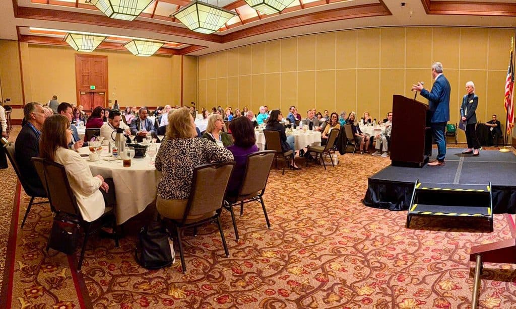 A ballroom of seated REALTOR attendees at banquet tables listening to a speaker at a podium on a low stage.