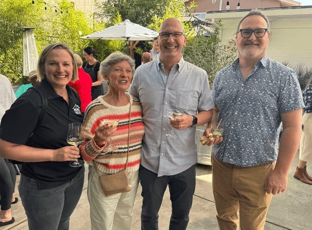 Four people holding wine and cocktail glasses at an outdoor patio gathering under string lights.