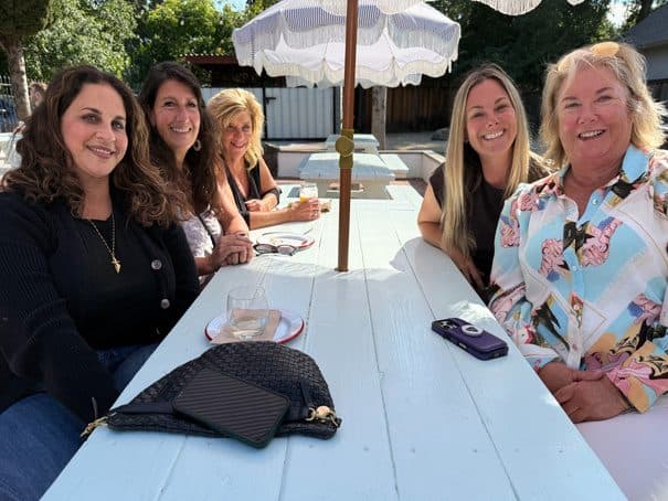 Five attendees smiling around a long white outdoor table under a patio umbrella at a summer chapter mixer.