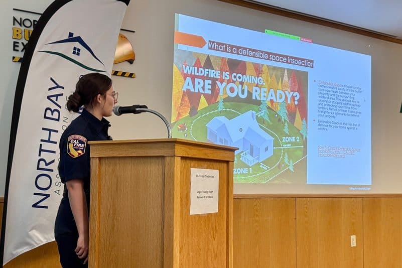A CAL FIRE presenter at the podium beside a slide asking "Wildfire is coming. Are you ready?" with defensible space zone diagrams.