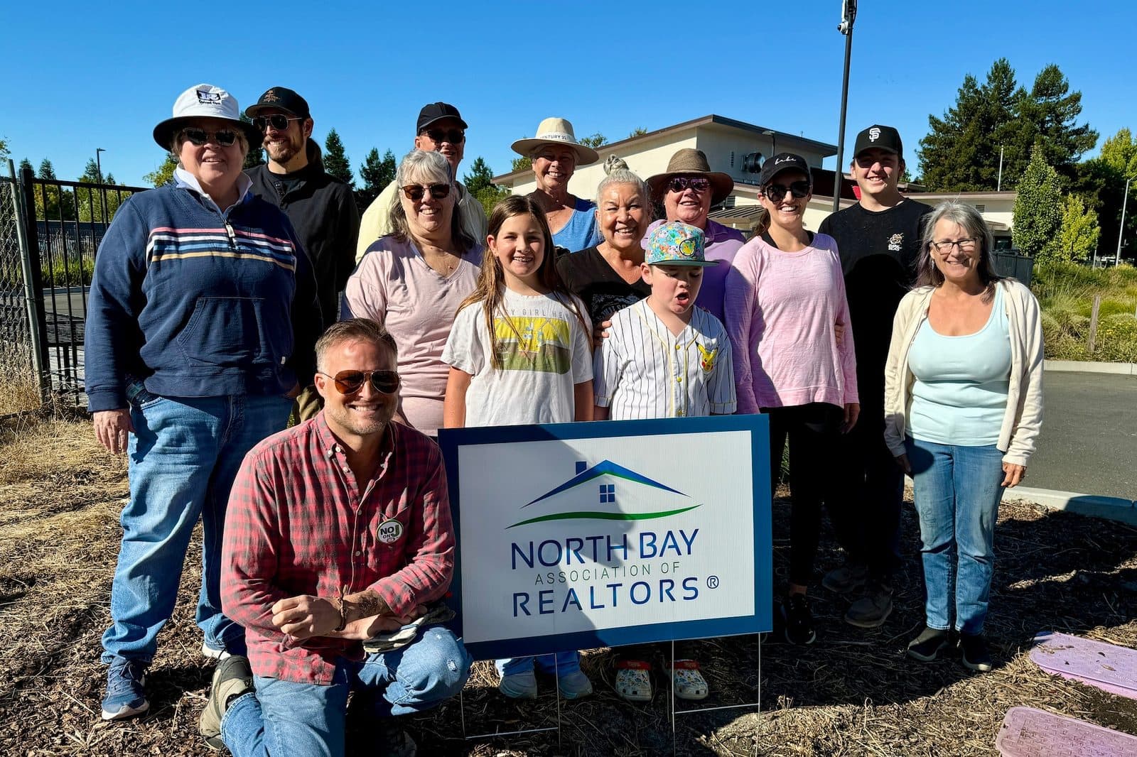 Group of volunteers including two children standing outdoors behind a North Bay Association of REALTORS yard sign at the creek clean-up.