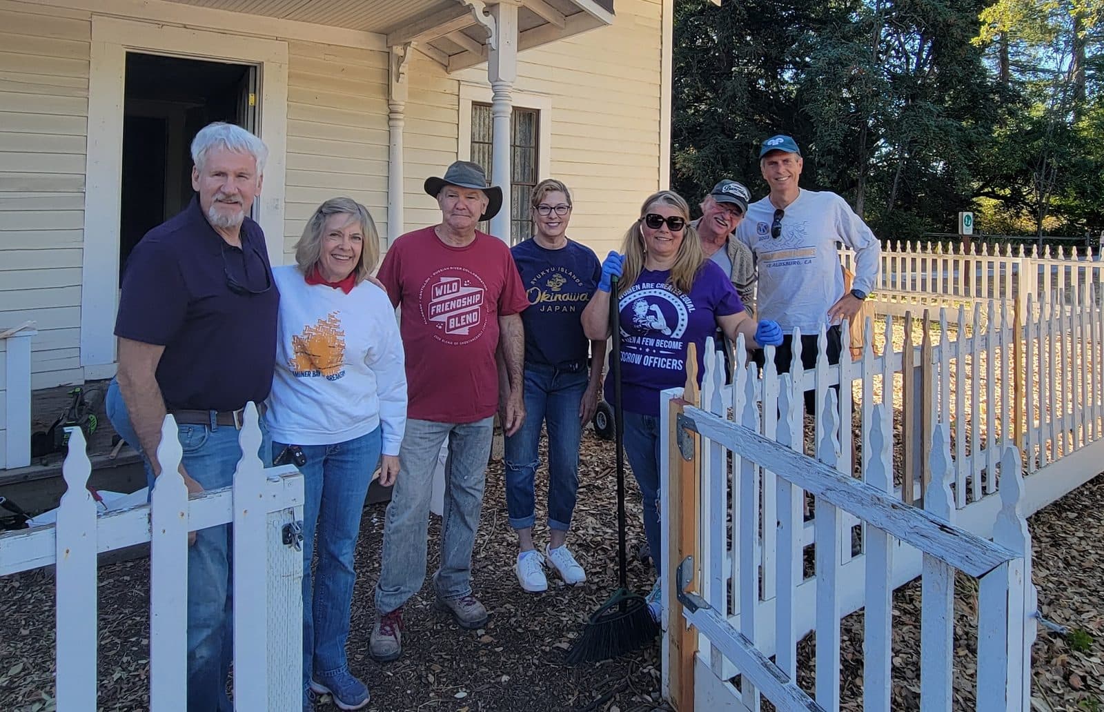 Seven volunteers posed at a freshly painted white picket fence in front of a historic clapboard house.