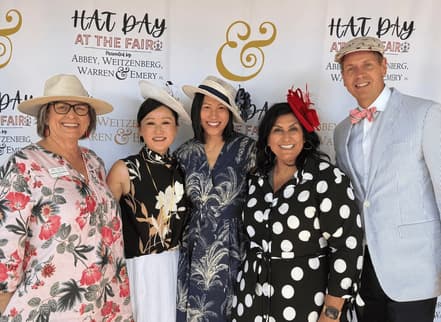 Six guests in summer hats and dresses posed in front of the Hat Day at the Fair backdrop.