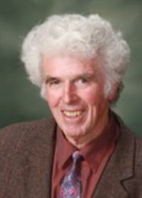 Stephen Kent Jones, smiling, with curly white hair, in a brown tweed jacket, red shirt and patterned tie.