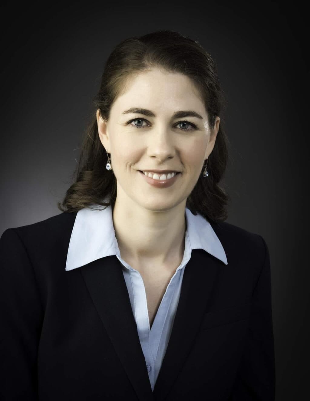 A woman with dark shoulder-length hair in a black blazer over a pale blue shirt, against a dark grey studio background.
