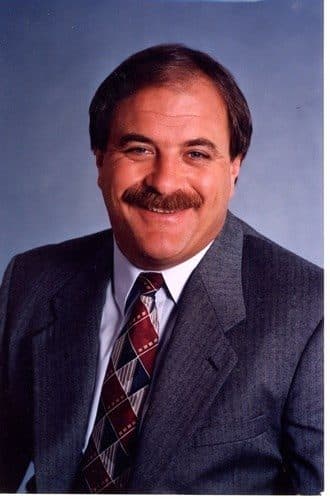Stephen Herndon, smiling, in a grey pinstripe suit and patterned burgundy tie against a blue-grey studio background.