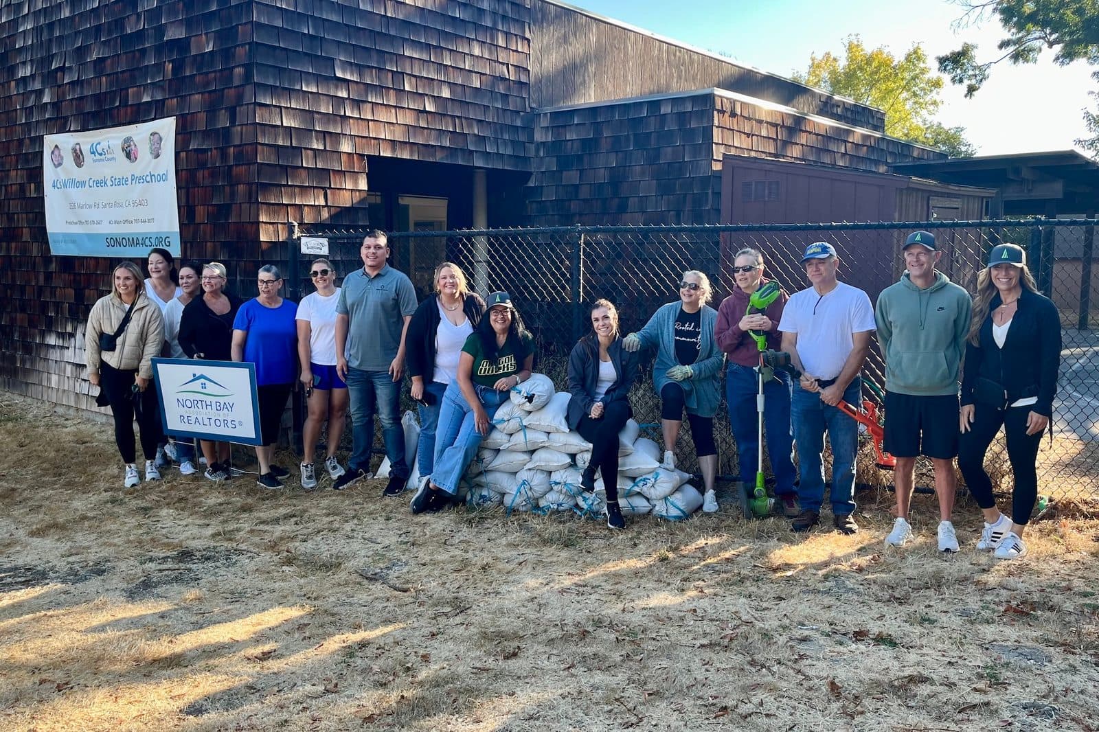 Around fifteen volunteers lined up in front of a shingled preschool building beside a stack of soil bags, with a North Bay Association of REALTORS yard sign at left and a 4Cs Willow Creek State Preschool banner behind them; two hold garden power tools.