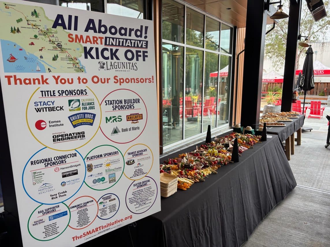 A large All Aboard! SMART Initiative Kick Off sponsor board listing sponsor logos, standing beside a long grey-skirted table laid with a charcuterie spread on a covered patio.