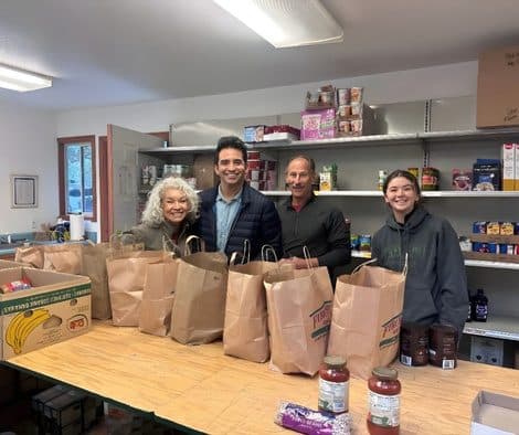 Four volunteers standing behind a long table stacked with filled paper grocery bags in a food pantry storeroom, shelves of canned goods behind them.